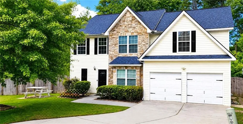Traditional two-story residence featuring a stone and siding facade, a dark blue shingle roof, a two-car garage, and established landscaping