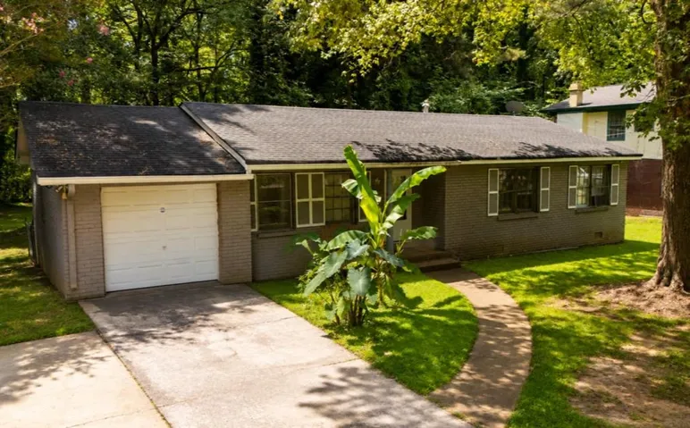 Exterior featuring a gray brick facade, attached garage, paved driveway, concrete walkway, and mature landscaping