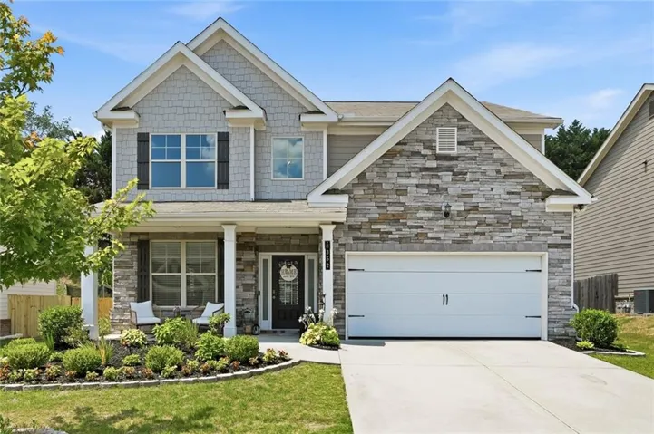 Two-story residence featuring a stone and shingle facade, a covered front porch with columns, a manicured front yard, and an attached two-car garage with a concrete driveway