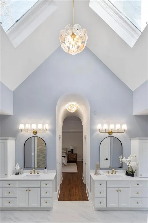 Ensuite bathroom featuring a chandelier, two vanities, a skylight, high vaulted ceiling, and light marble finish floors