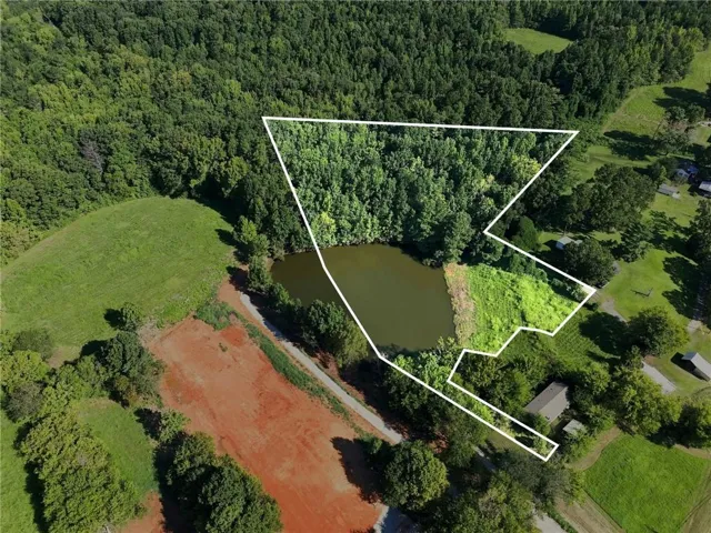Aerial view of an expansive wooded property featuring a natural pond bordered by mature forest, open green pasture, and cleared land