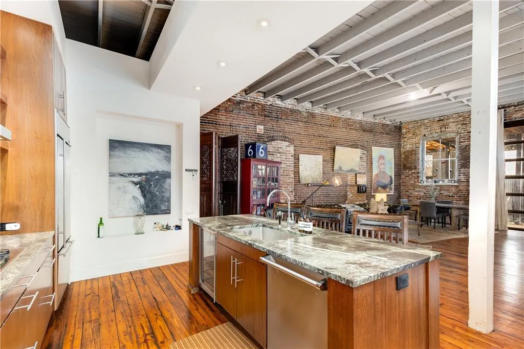 Kitchen with brick wall, brown cabinetry, dark wood-type flooring, stainless steel dishwasher, and light stone countertops