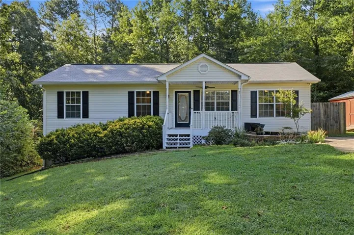 Exterior featuring light-colored siding, dark window shutters, a covered front porch with white railings, and a landscaped front yard with established plantings