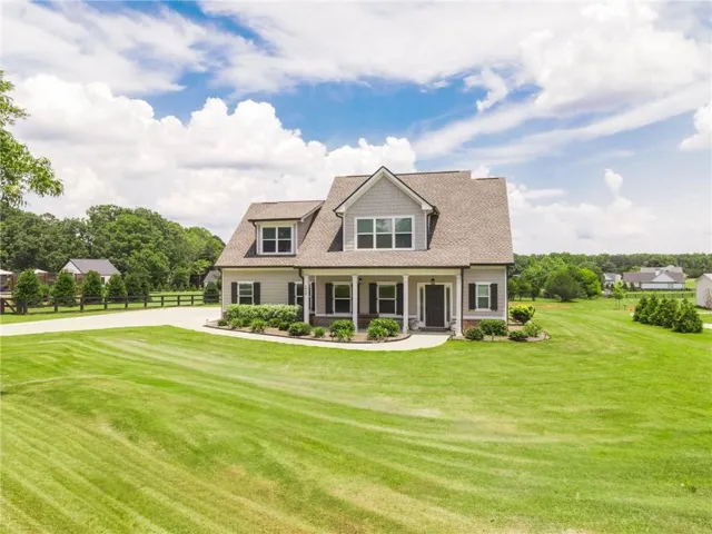 Two-story residence featuring light gray siding, dark shutters, a covered front porch, and dormer windows, surrounded by a expansive green lawn and mature landscaping