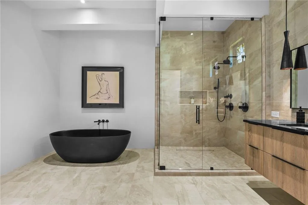 Freestanding matte black soaking tub, glass-enclosed shower with tiled walls and built-in niche, dual shower heads, floating wood-finish vanity with black countertop, and coordinated pendant lighting