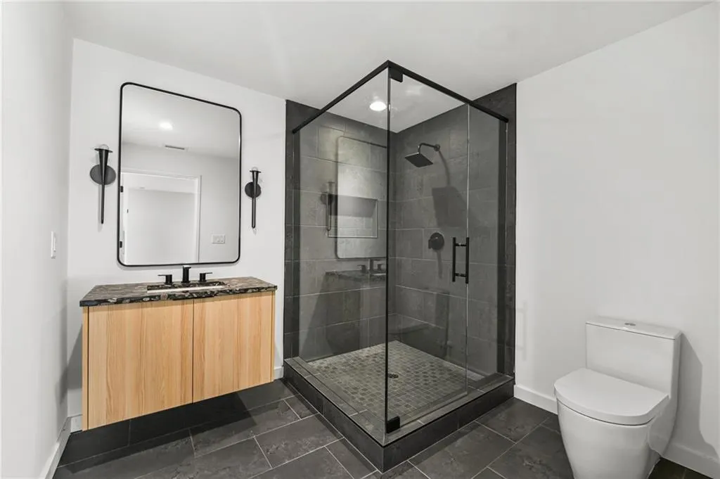 Floating wood-finish vanity with a dark countertop, framed mirror, and a glass-enclosed shower featuring dark tile