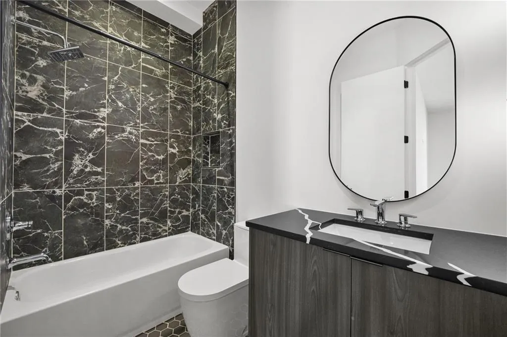 Black marble-style wall tiling, white bathtub with a built-in shower niche, dark wood-finish vanity with an integrated sink, and an oval-shaped mirror with a black frame