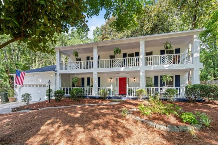 Two-story columned facade, double-level balconies with decorative railings, vibrant red entry door, attached two-car garage, and mature landscaping