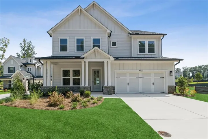 Two-story residence featuring light-colored siding, a covered front porch with columns, a two-car garage, stone accents, and a concrete driveway