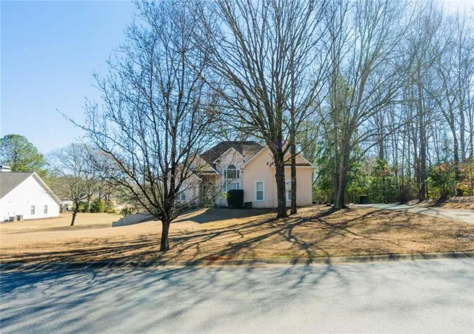 Spacious lot featuring a light-colored exterior residence with a dark roof, mature trees, and a paved street