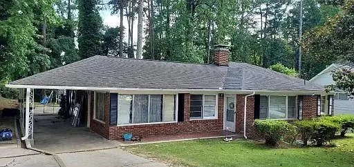 Brick exterior featuring a covered carport, full-length windows, a prominent brick chimney, and mature trees surrounding the property
