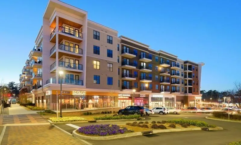 Modern multi-story building featuring ground-level retail spaces, residential balconies with glass railings, and a landscaped streetscape