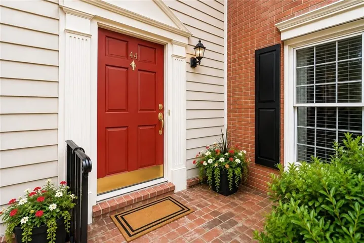 Red paneled entry door with brass hardware, brick paver porch, and white siding