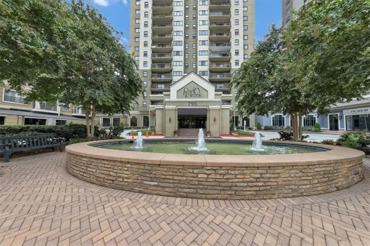 Multi-story building exterior with individual balconies, a stone-clad fountain, herringbone paver hardscaping, and mature trees