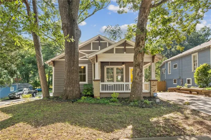 Craftsman-style exterior with gabled roofline, front porch with white railing, brick base columns, and mature trees providing natural shade