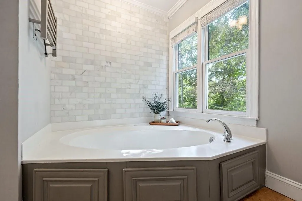 Corner soaking tub featuring a stone-tile backsplash, white surround, and grey custom cabinetry base, complemented by a large double-hung window