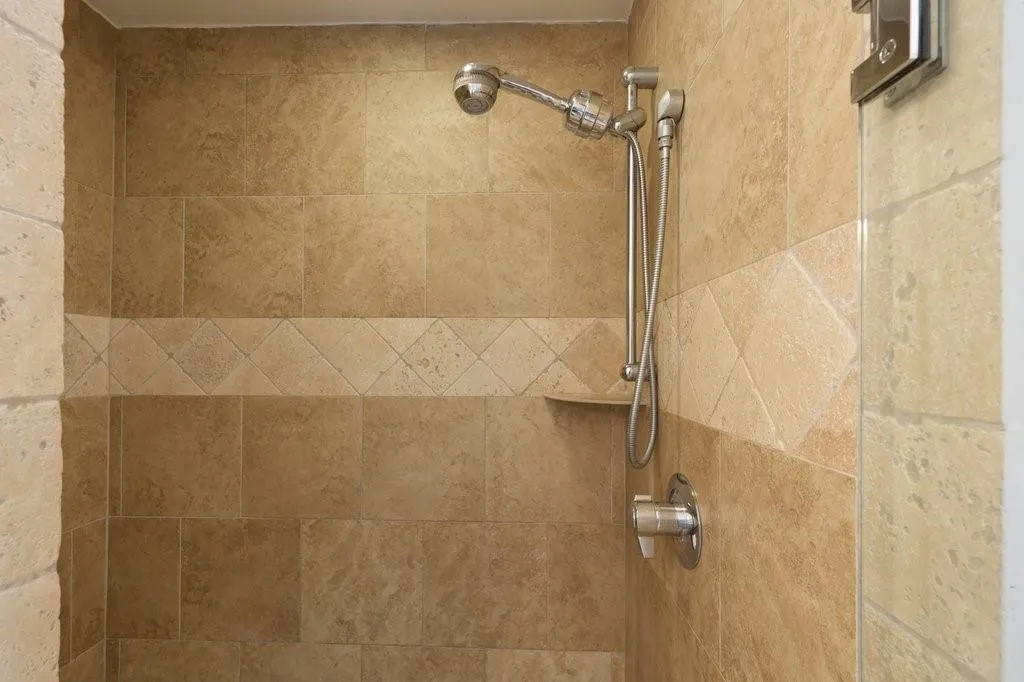 Walk-in shower featuring floor-to-ceiling stone-finish wall tiles, a diagonal-pattern tile inlay, brushed-metal adjustable shower head with handheld attachment, built-in corner shelf, and a glass partition