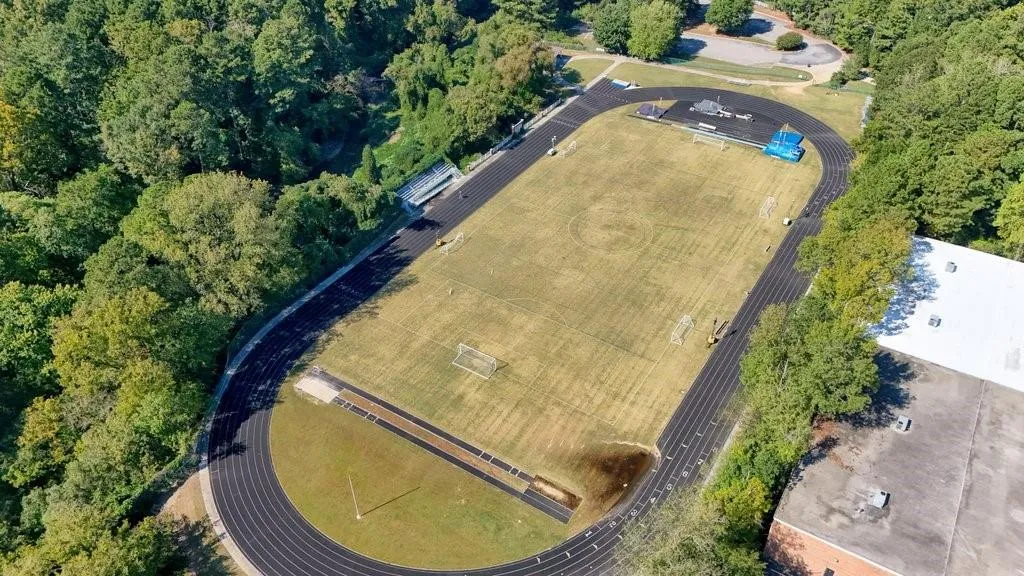 Aerial perspective of a multi-purpose athletic field featuring a paved running track, grass turf, and perimeter metal bleachers