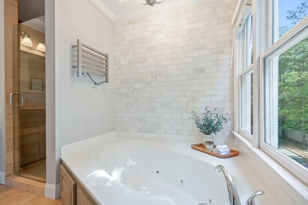 Jetted soaking tub with chrome fixtures, floor-to-ceiling brick-pattern stone tile surround, wall-mounted electric towel warmer, glass-enclosed shower, and large multi-pane windows