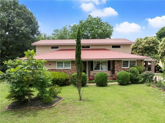 Red metal roof, brick facade, covered porch, mature landscaping, and an expansive lawn