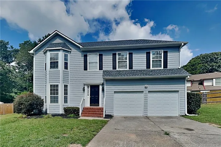 Two-story facade with light-colored siding, contrasting dark shutters, a prominent bay window, an attached two-car garage, and a concrete driveway