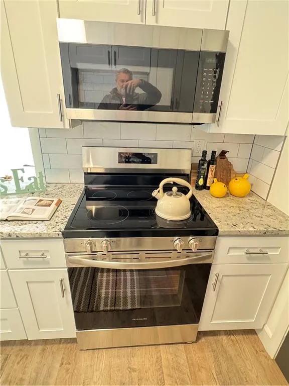 Kitchen with backsplash, white cabinets, stainless steel stove, light wood-type flooring, and light stone counters