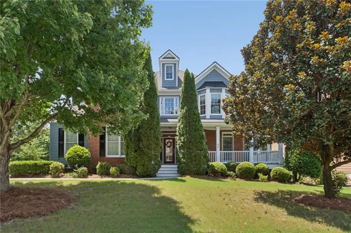 Two-story residence featuring a bay window, a columned front porch with white railings, a dormer window, and mature trees framing the property