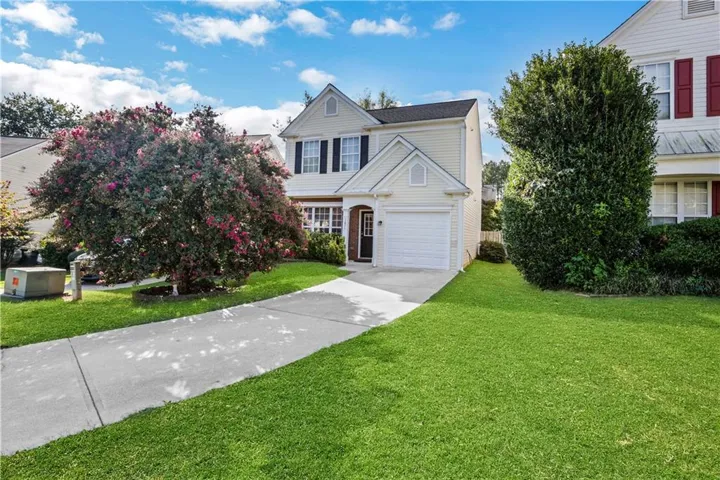 Light-colored siding exterior, attached garage, concrete driveway, manicured lawn, and mature flowering tree