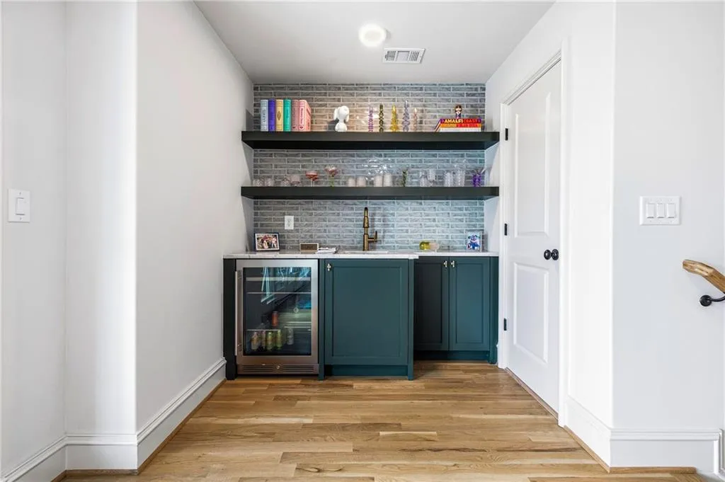 Built-in wet bar featuring teal cabinetry, a beverage refrigerator, a sink with a brass-finish faucet, and two floating wood shelves set against a light brick-patterned tile backsplash