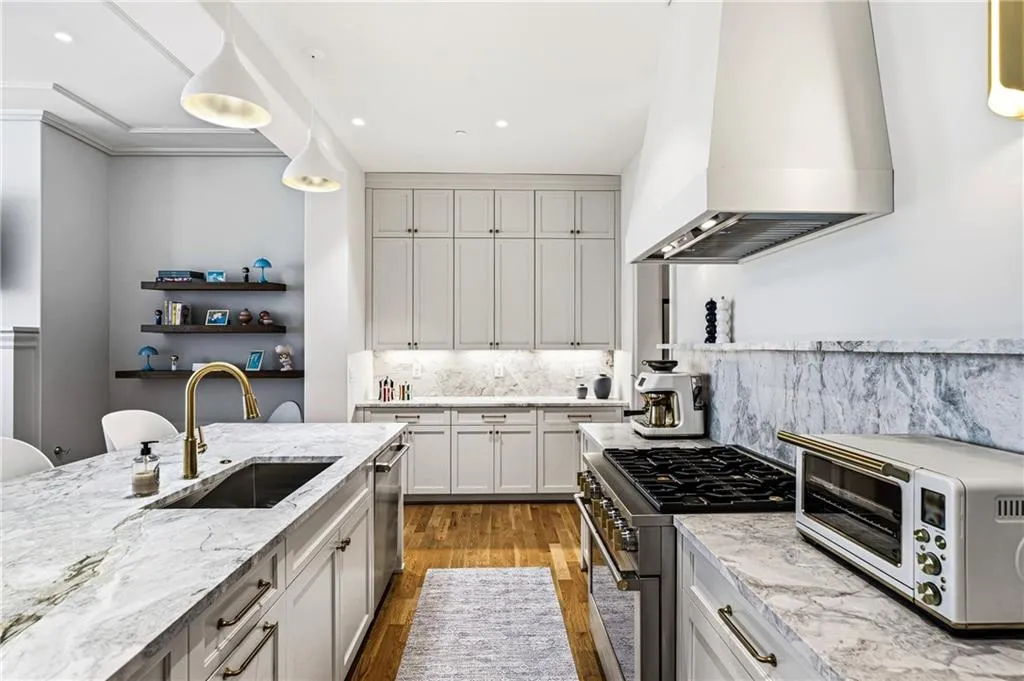 Kitchen featuring a large island with an undermount sink and brass-finish faucet, extensive light-toned cabinetry, stainless steel range with a professional-grade hood, and light-toned stone countertops with matching backsplash