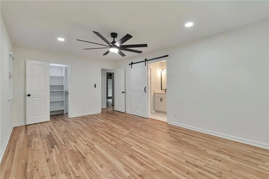 Unfurnished bedroom featuring light wood-type flooring, ensuite bath, ceiling fan, a walk in closet, and a barn door