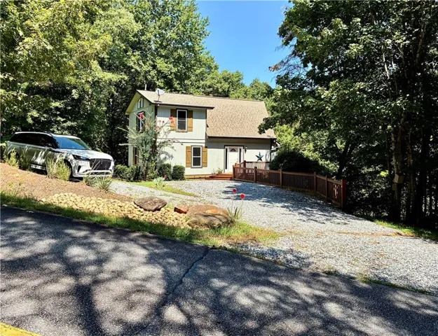 Two-story residence with a light-colored exterior, a gravel driveway, wood-toned shutters, and a wood-toned railing leading to the front entry
