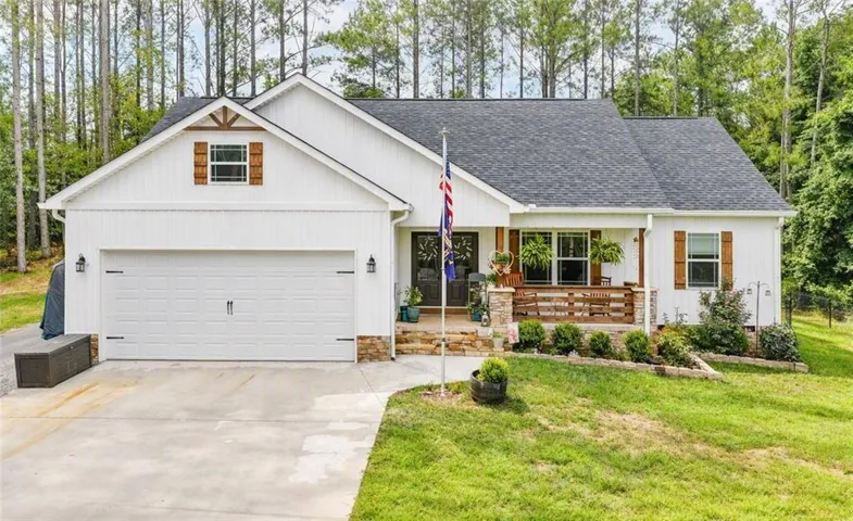 White vertical siding with dark shingles, an attached garage, and a covered front porch featuring stone accents and wooden railings