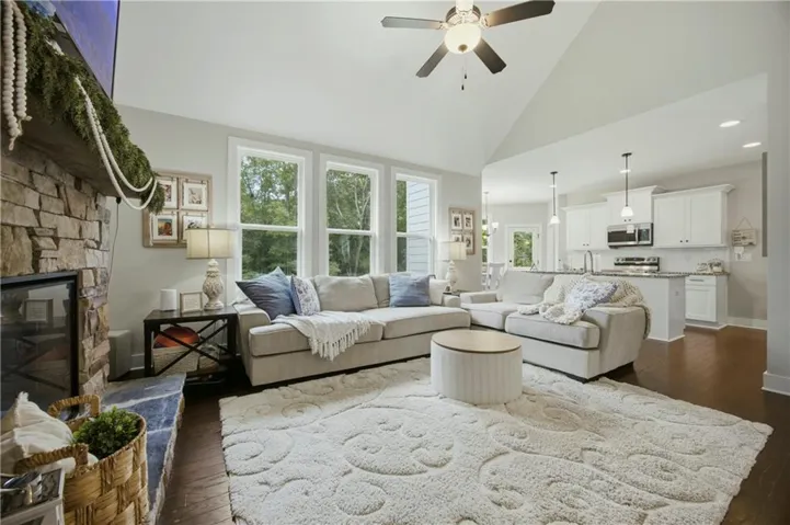 Spacious living area featuring a stone-clad fireplace, vaulted ceiling with ceiling fan, wood-finish flooring, and multiple windows