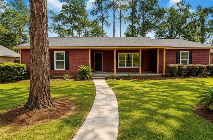 Red brick single-story residence featuring a covered front porch with wood-finish columns, manicured lawn with mature pine trees, curved concrete walkway, and lush landscaping with ornamental plantings throughout the grounds