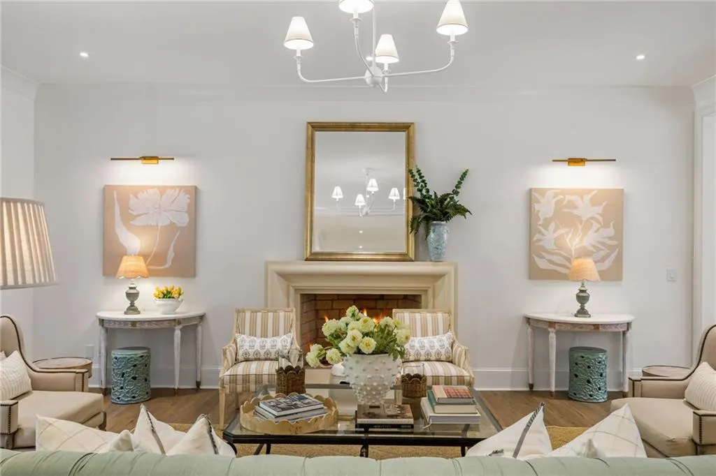 Fireplace with an architectural stone-style mantel, warm-toned wood-finish flooring, white five-arm chandelier, and symmetrical wall-mounted gold-toned sconces