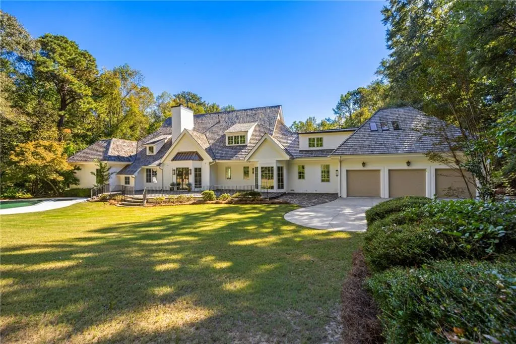 Custom residence featuring a shingled roof, white exterior cladding, three-car garage, manicured lawn, and mature perimeter trees