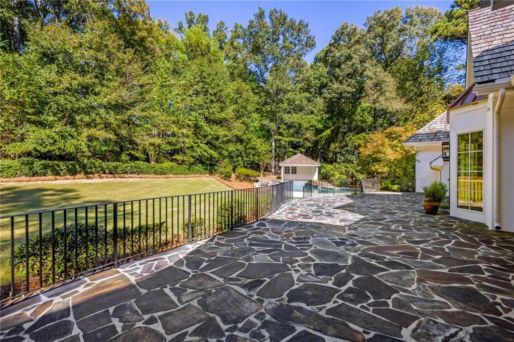 Flagstone patio with metal perimeter fencing, expansive lawn, mature tree line, and an in-ground swimming pool