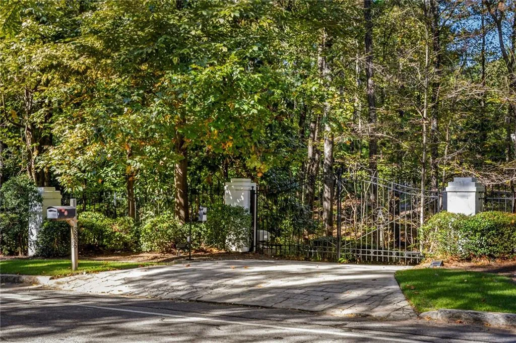 Private entrance featuring a paved stone driveway, decorative wrought-iron gate, and concrete pillars set among mature trees and landscaping