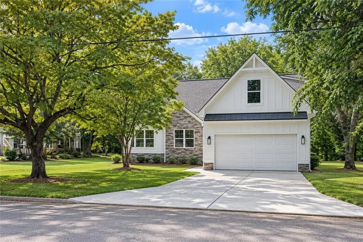 Gabled white board-and-batten and stone facade with a two-car garage, concrete driveway, and mature landscaping