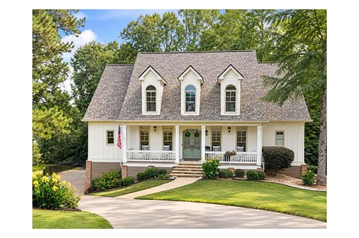 Dormered roofline with arched windows, white siding, covered front porch, brick foundation, and a verdant lawn