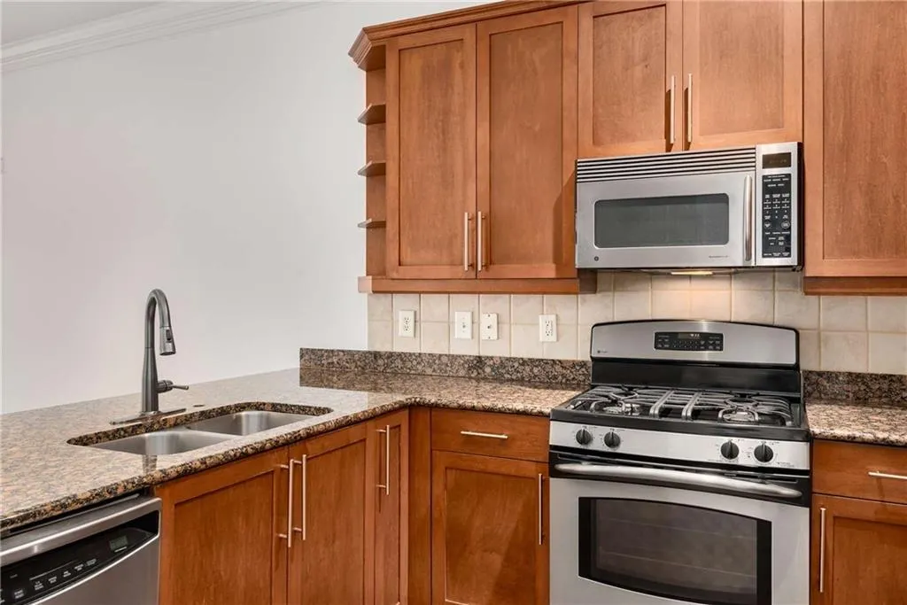 Kitchen featuring stainless steel appliances, open shelves, brown cabinets, backsplash, and dark stone counters