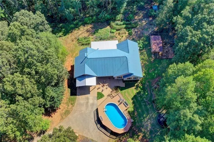Aerial perspective showcasing a blue metal roof, an oval-shaped pool with surrounding decking, a concrete driveway, a covered patio, and abundant mature trees