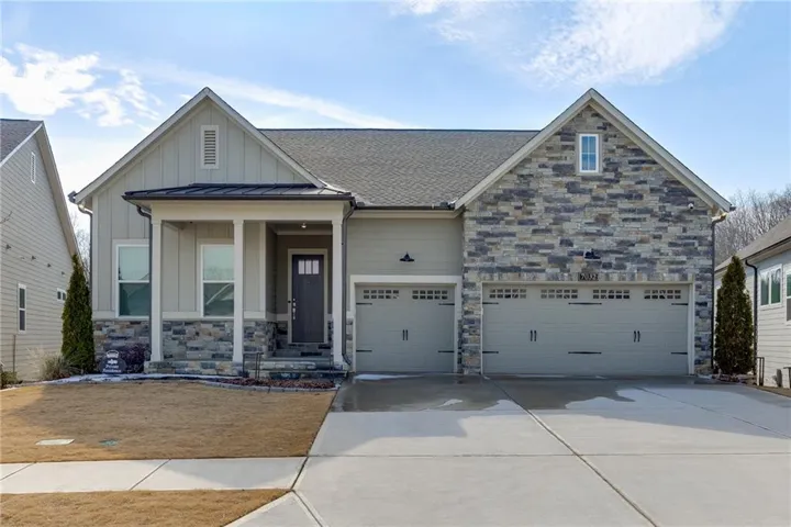 Modern brick and stone exterior featuring a covered front porch with substantial columns, dual three-car garage with paneled doors, manicured front lawn with mature evergreen landscaping, and a wide concrete driveway