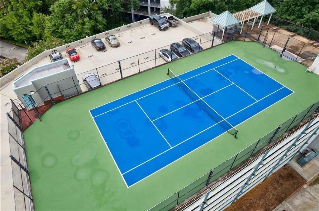 Outdoor tennis court with blue and green surfacing, perimeter fencing, and an adjacent parking area