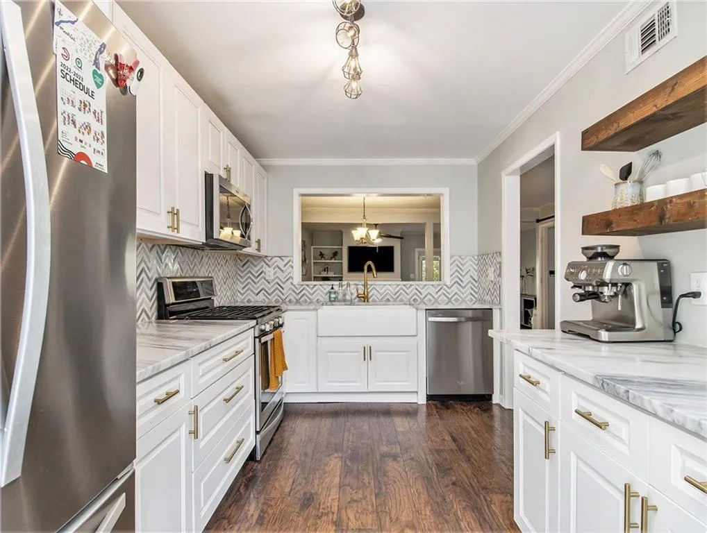 Kitchen featuring wood-finish flooring, white cabinetry with brass hardware, a white farmhouse sink with a gold-tone faucet, stainless steel appliances, and open wood shelving