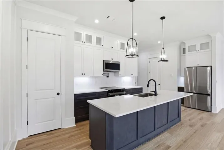 Modern kitchen featuring a central island with a white countertop and black base, wood-finish flooring, white upper cabinetry, stainless steel appliances, and two pendant light fixtures