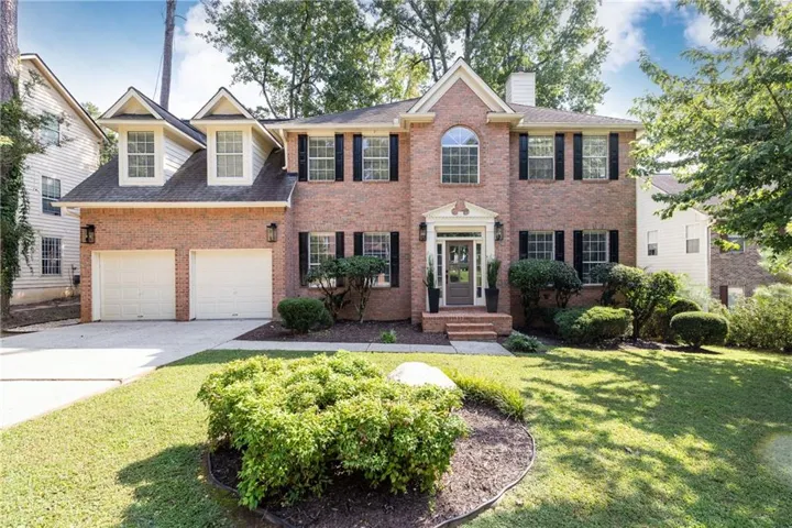 Brick facade with prominent white trim, black louvered shutters, gabled dormers, a two-car garage, and a concrete driveway