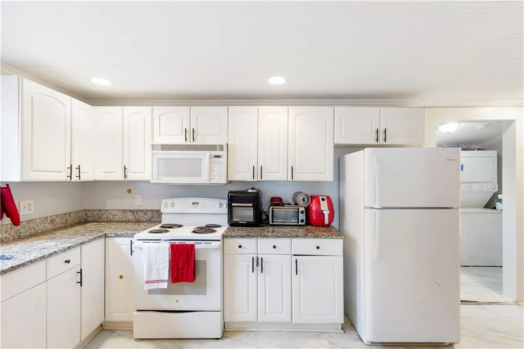 Kitchen with light tile floors, white cabinetry, light stone countertops, white appliances, and stacked washer and dryer