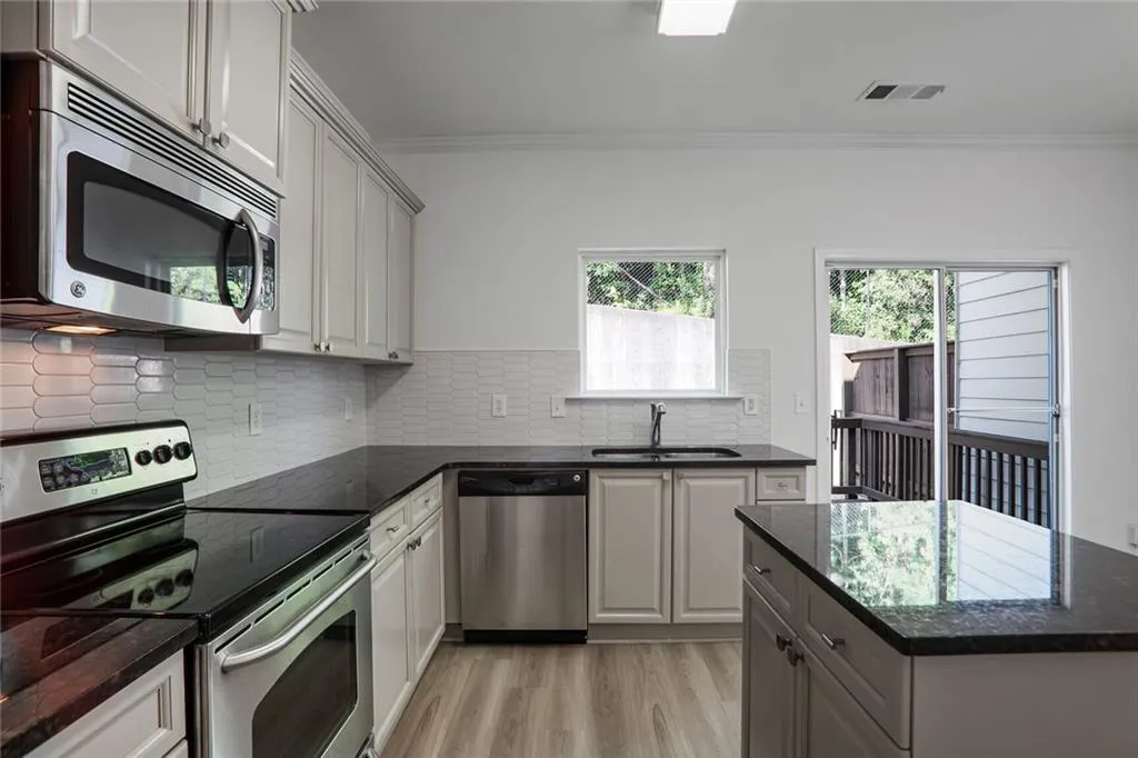 Kitchen with appliances with stainless steel finishes, crown molding, light wood finished floors, decorative backsplash, and dark stone countertops